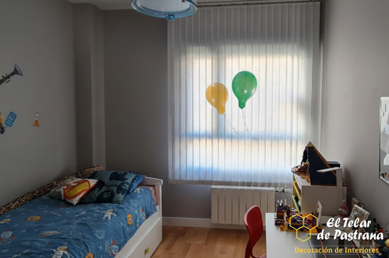 Habitación infantil: cortina vertical blanca en ventana sobre radiador, cama individual con ropa de cama azul, paredes grises y dos globos flotando cerca de la cortina.