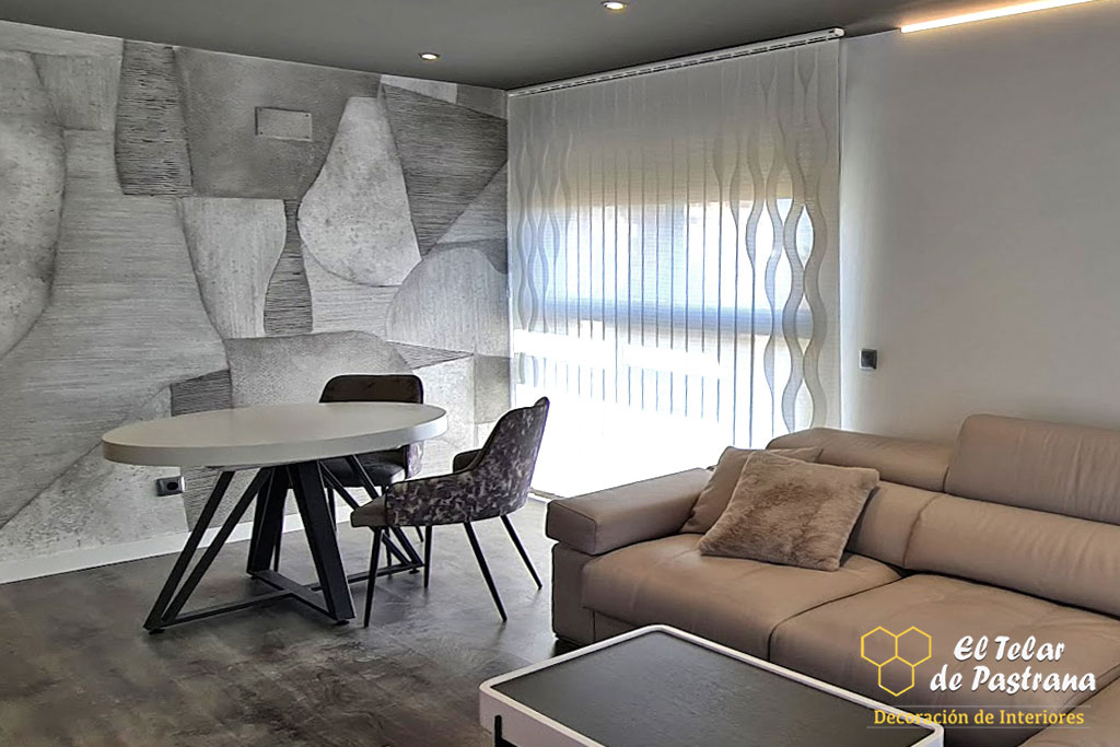 Sala de estar: cortina vertical blanca con diseño de ondas en ventana, sofá de piel beige, mesa de comedor redonda con sillas y pared con mural abstracto gris.