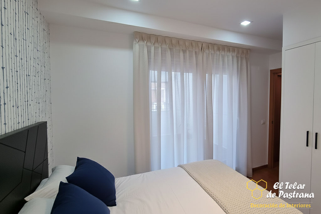 Dormitorio minimalista con pared en color topo, cama de madera con almacenaje y visillo de gran caída con pinza pliegue.