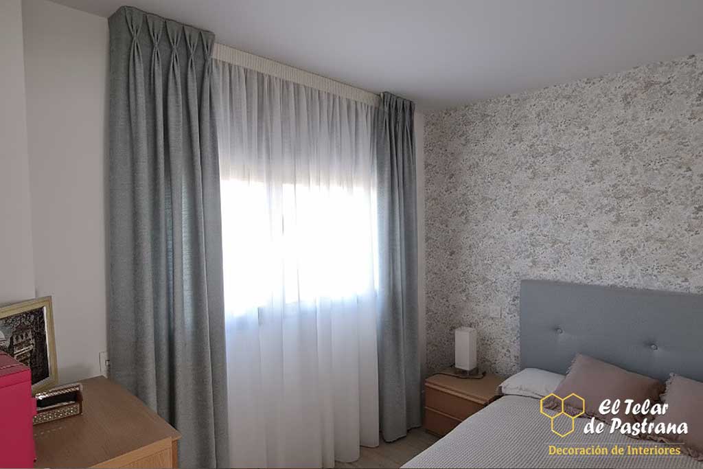 Cortinas de pinza pliegue grises y visillo blanco en un dormitorio de matrimonio con cómoda y mesita de noche de madera.