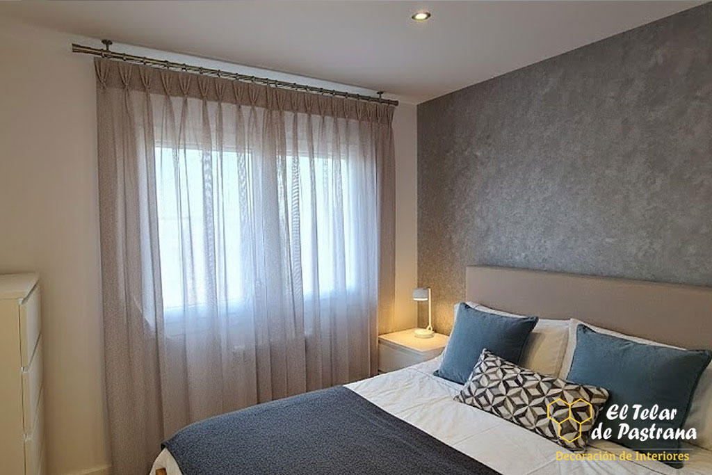 Dormitorio moderno con visillo de pinza pliegue en barra de metal, cama con cabecero beige y pared de fondo con textura gris.