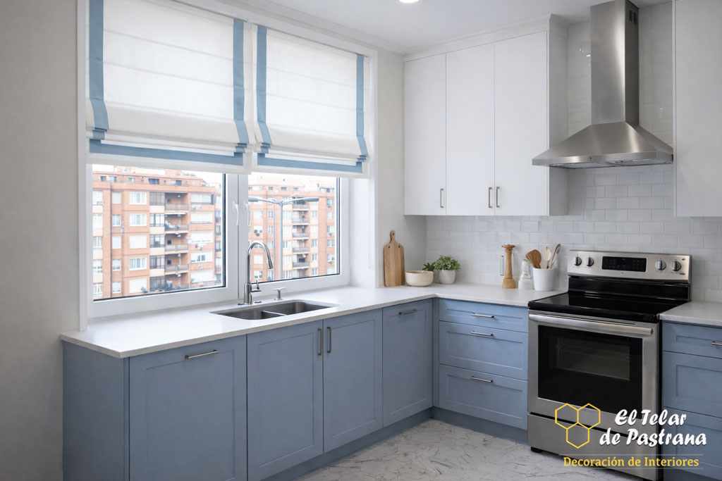 Cocina luminosa con muebles blancos y azules, estores plegables decorativos y zona de fregadero junto a la ventana